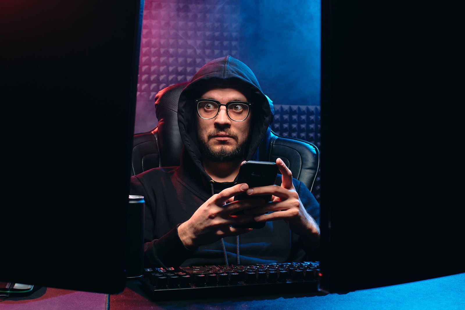 A man in a hoodie looking at his phone between two screens, in the dark